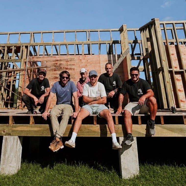 Six members of the team sitting on the floor platform of a timber house under construction, with the framing behind them.