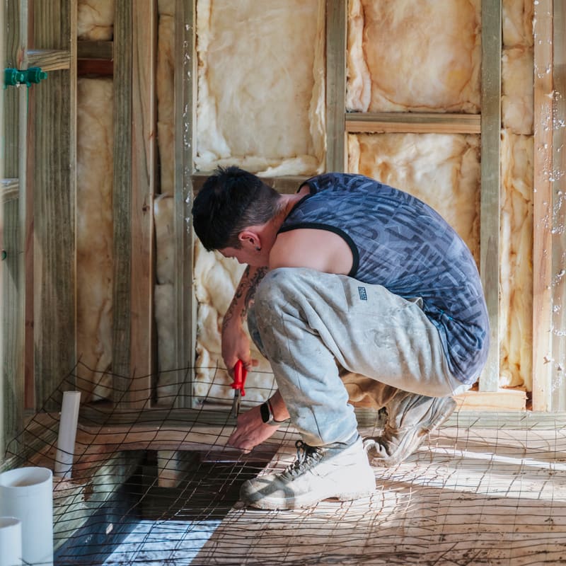 A builder works crouched on the floor between the insulated walls of a timber house.