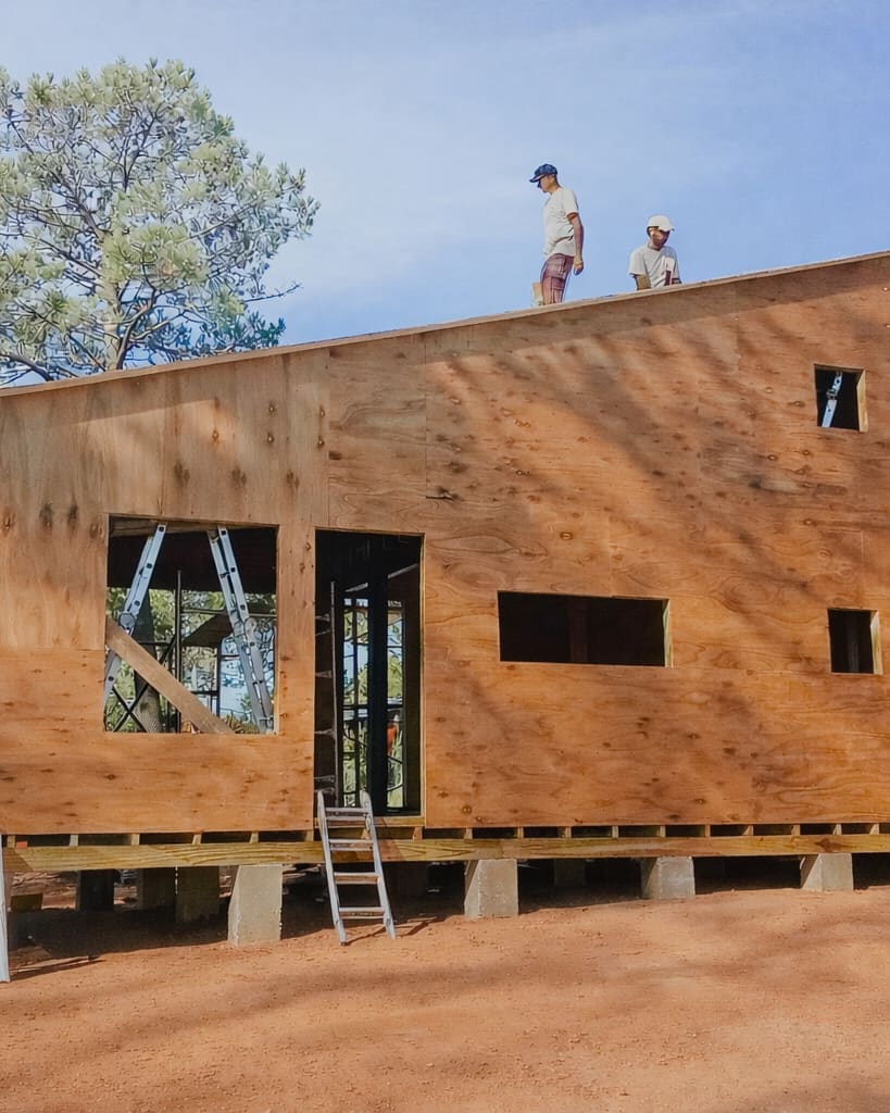 The house with its walls closed in panels, windows and doors not fitted yet. Three builders work on the scaffold and on the roof.
