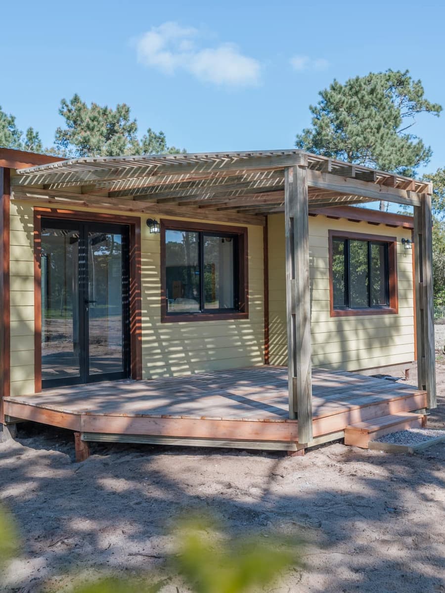 Finished timber house with a light facade and a covered wooden deck among pine trees.