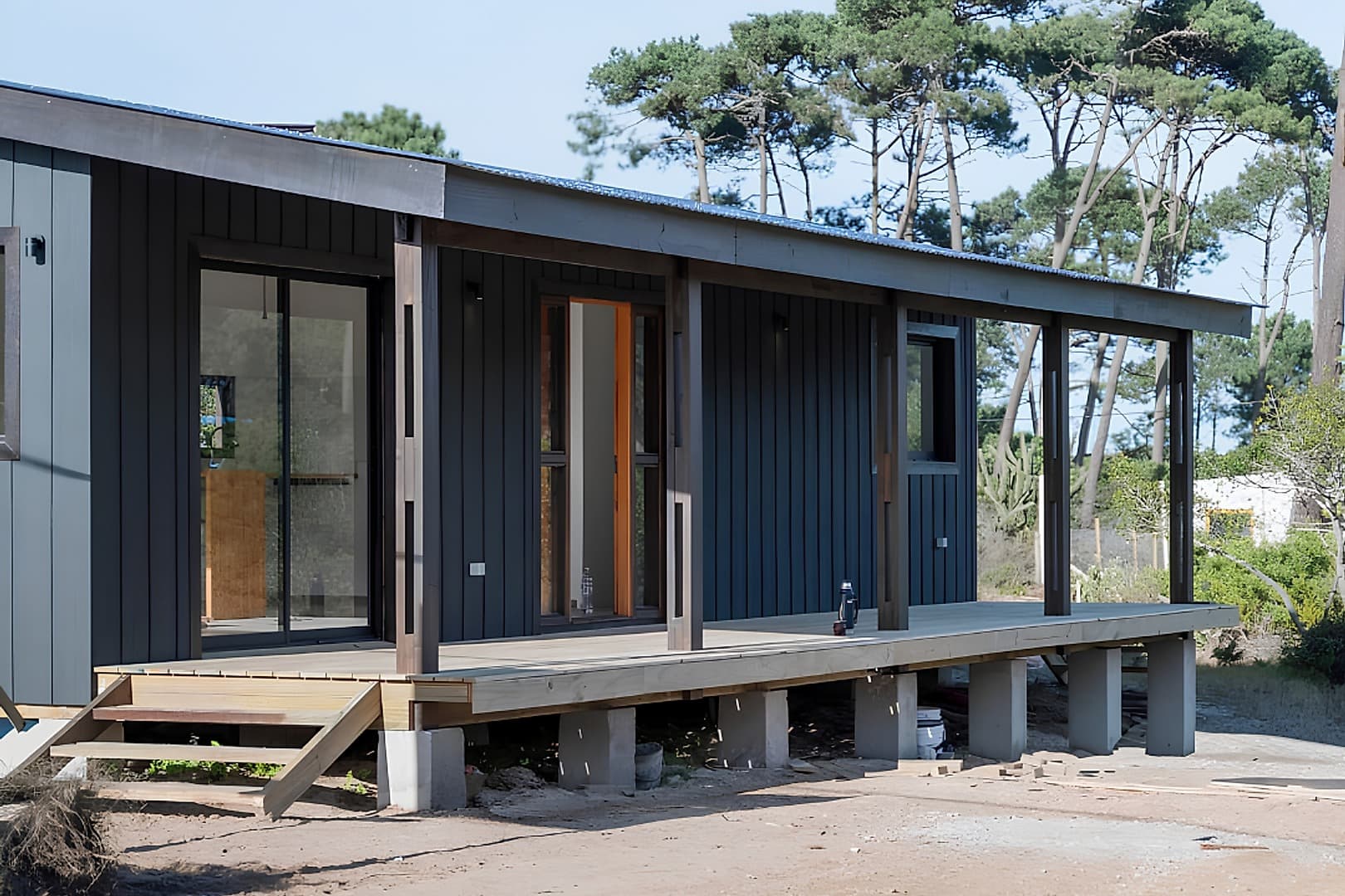 Completed TRES4CINCO timber house with dark vertical timber cladding, a covered gallery and a deck on concrete piers; the building layers are marked.