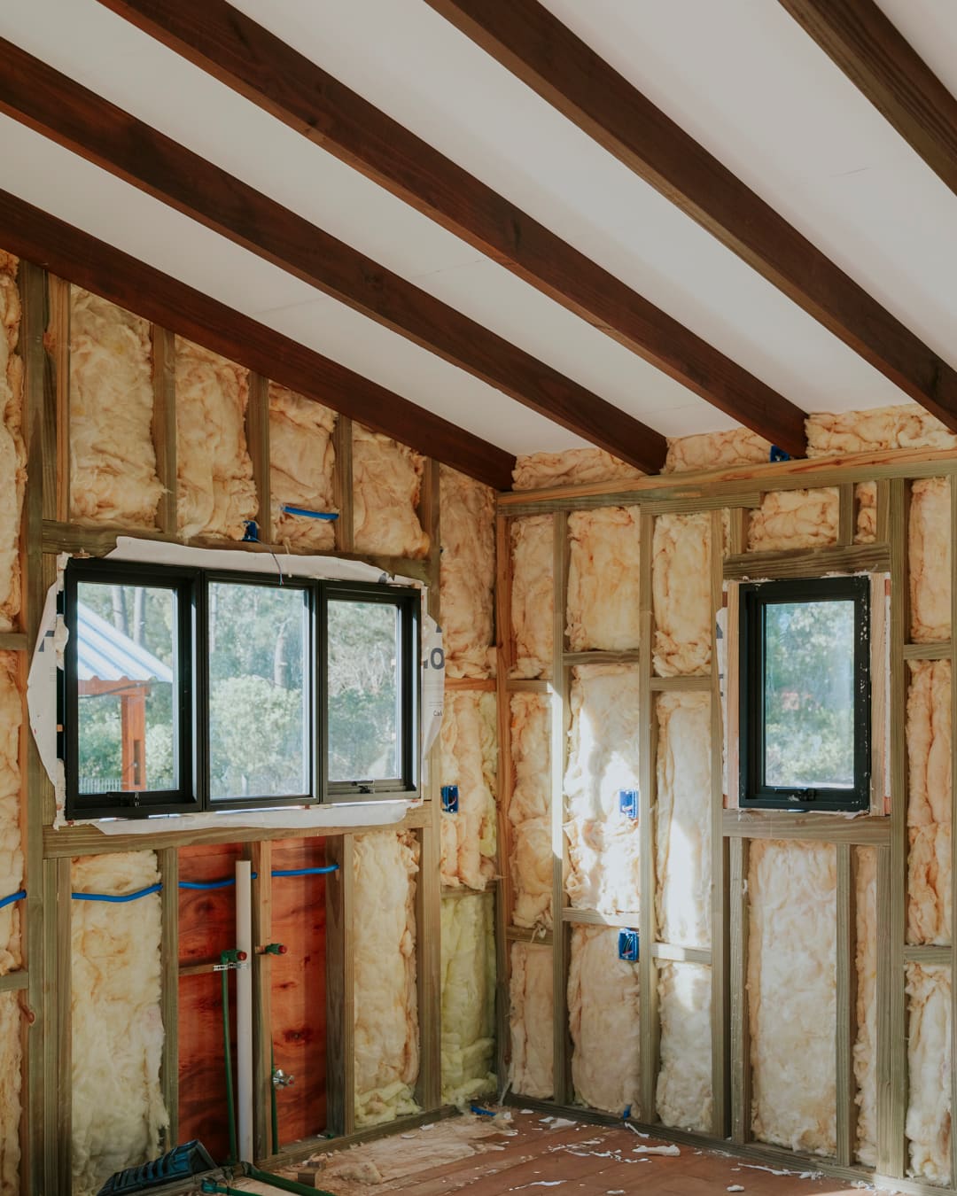 Corner of a room with mineral wool between the studs, two windows fitted, dark roof beams above.