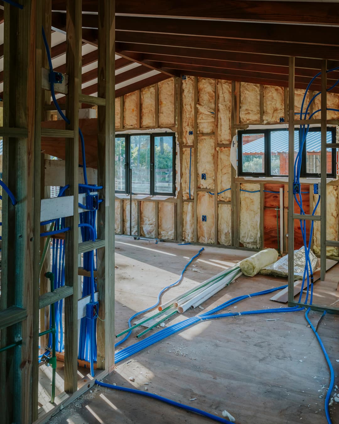 View through an open stud wall with blue conduits and water pipes into the room behind.
