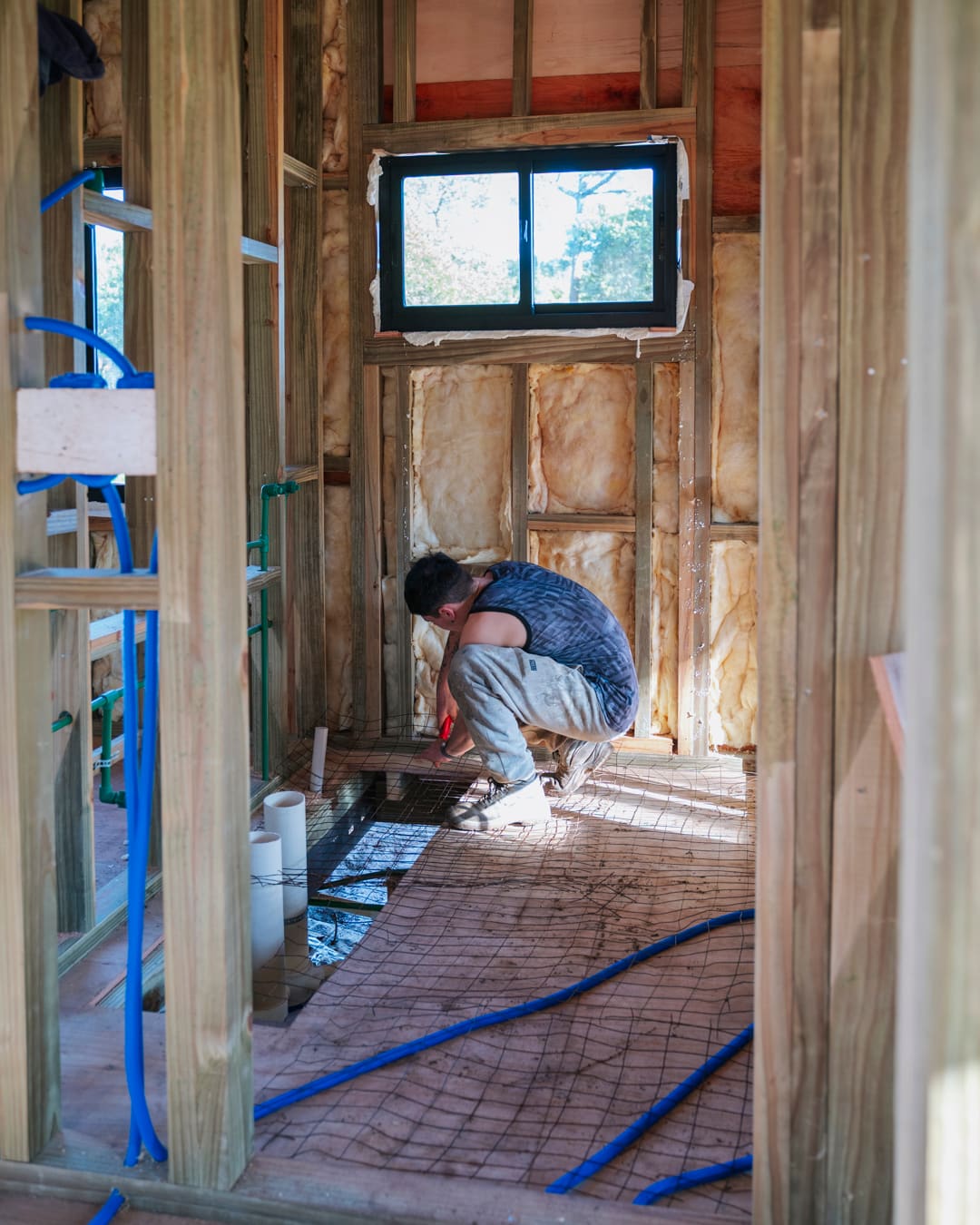 Worker kneeling on the floor of a small room between open studs, a small window above.