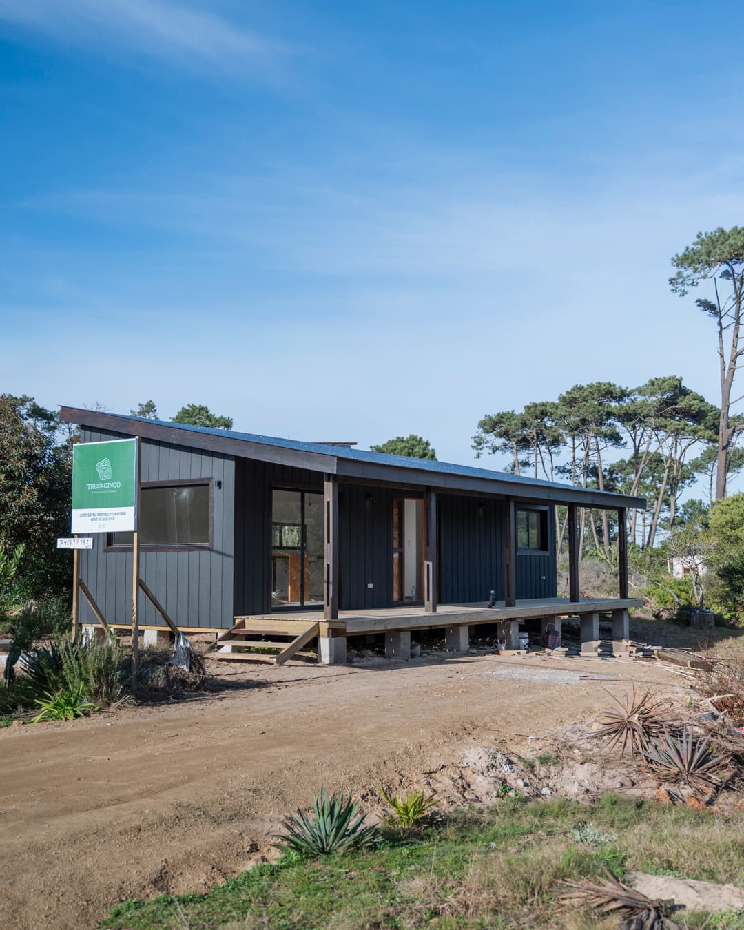 Long side of La Peregrina in daylight: black timber facade with veranda, the site sign beside it, pines in the background.