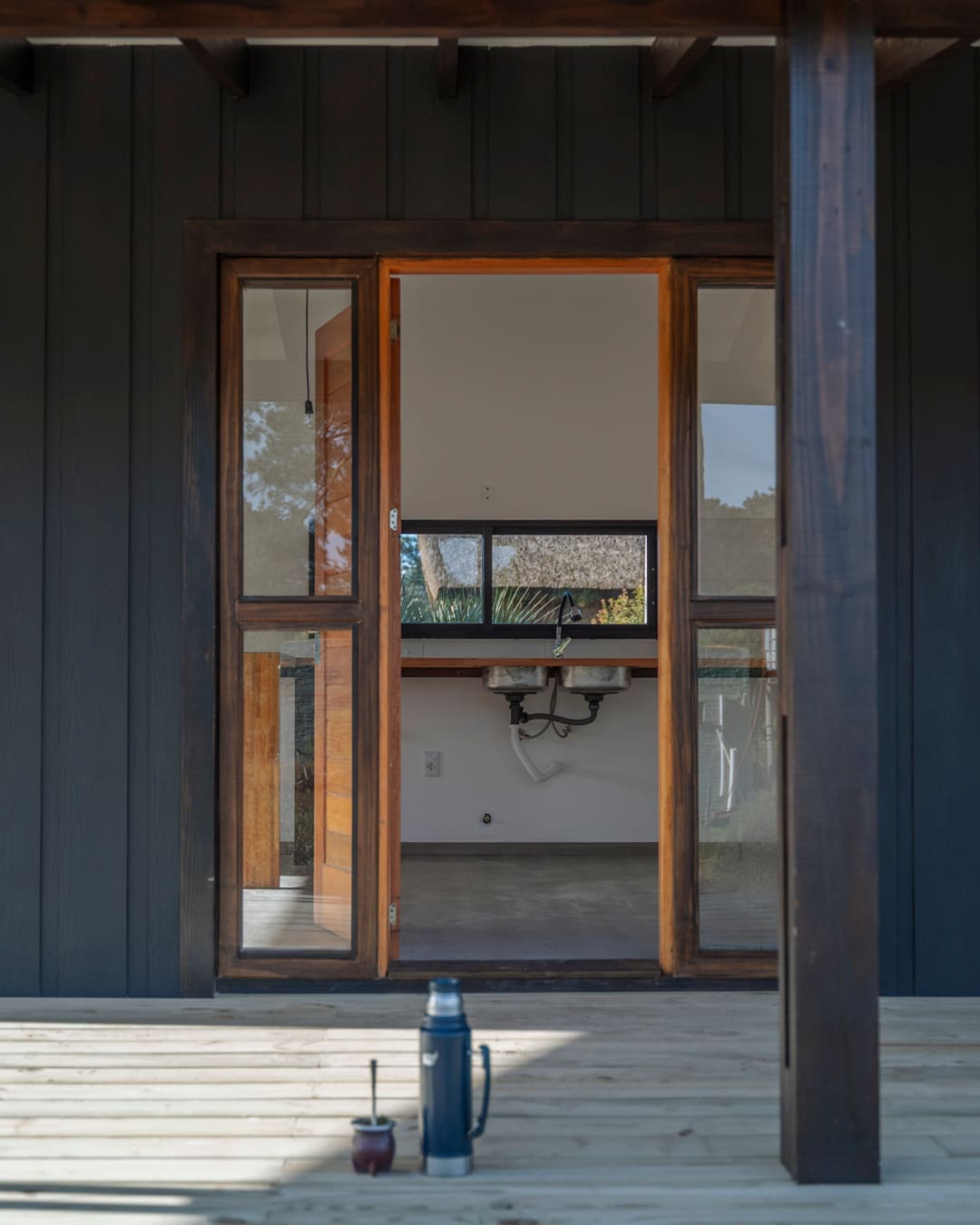 Timber front door with glazed panels standing open, view from the veranda into the kitchen; a mate and a thermos on the deck.