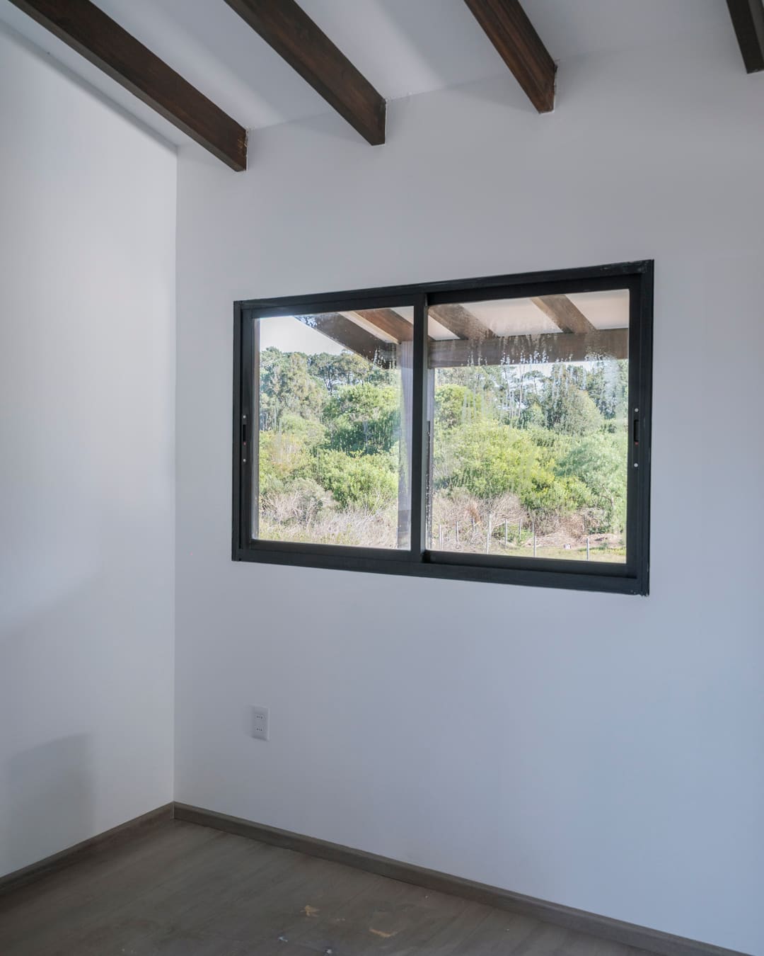 Room with a sliding window, white walls and dark roof beams, green outside.