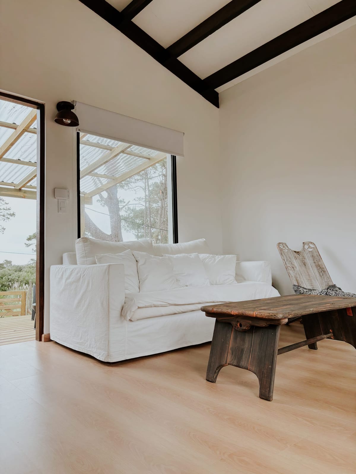 White sofa and rustic timber table under a high ceiling with dark beams, door and window to the terrace.