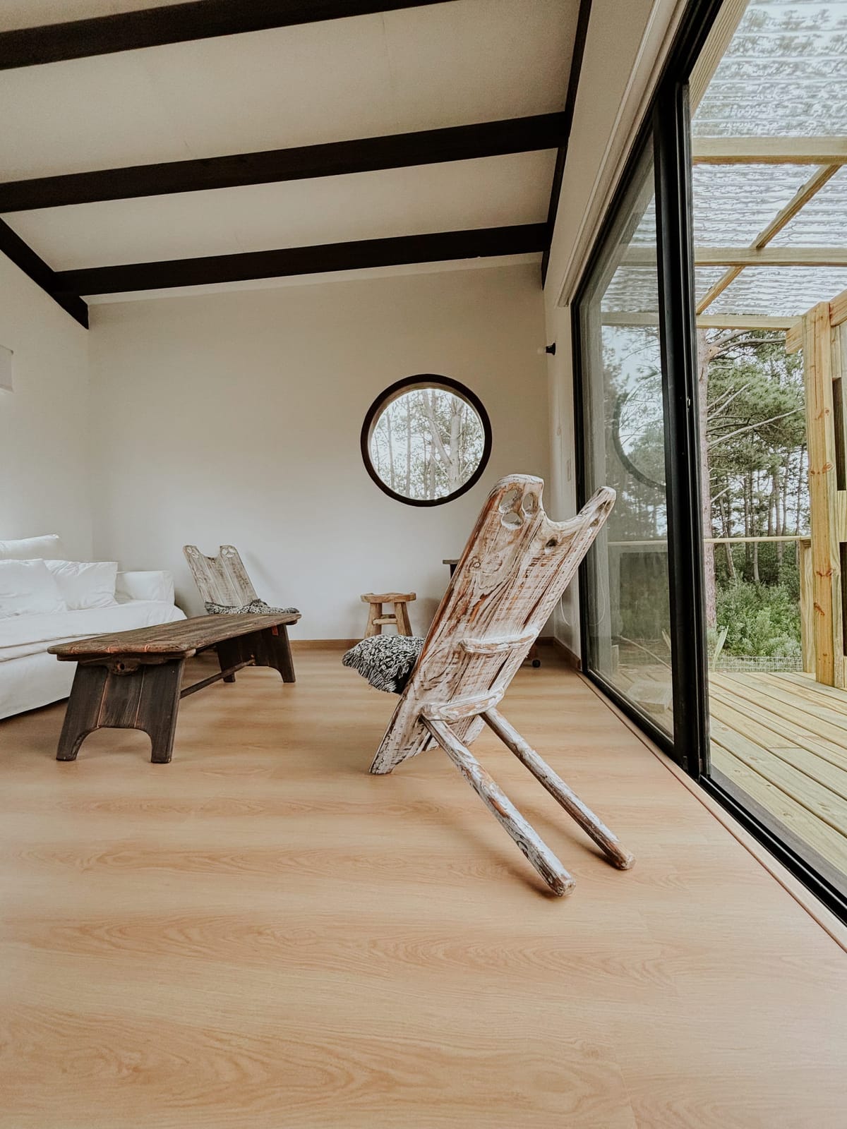 Living area of Matipipe with a rustic timber chair, round porthole window and open terrace door to the pine forest.
