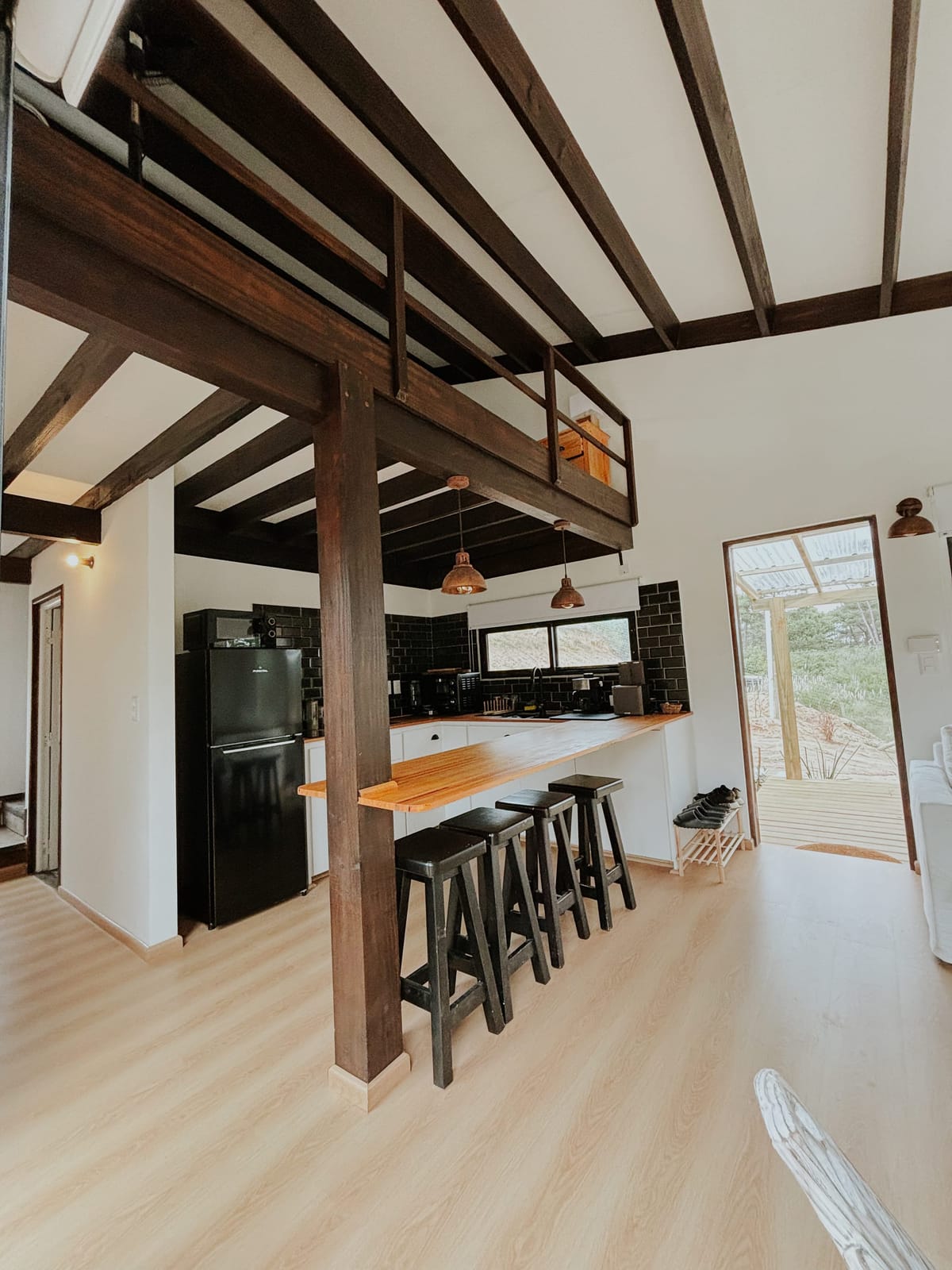 Open living space with kitchen counter and bar stools, the loft above on dark timber beams, the terrace door standing open.