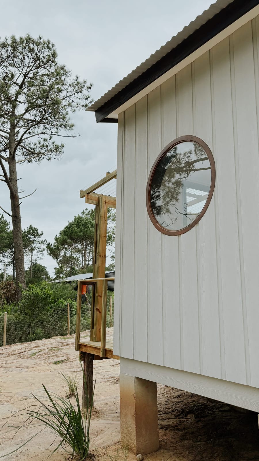 Side facade with a round porthole window and white timber cladding, the house raised on piers above the sand.