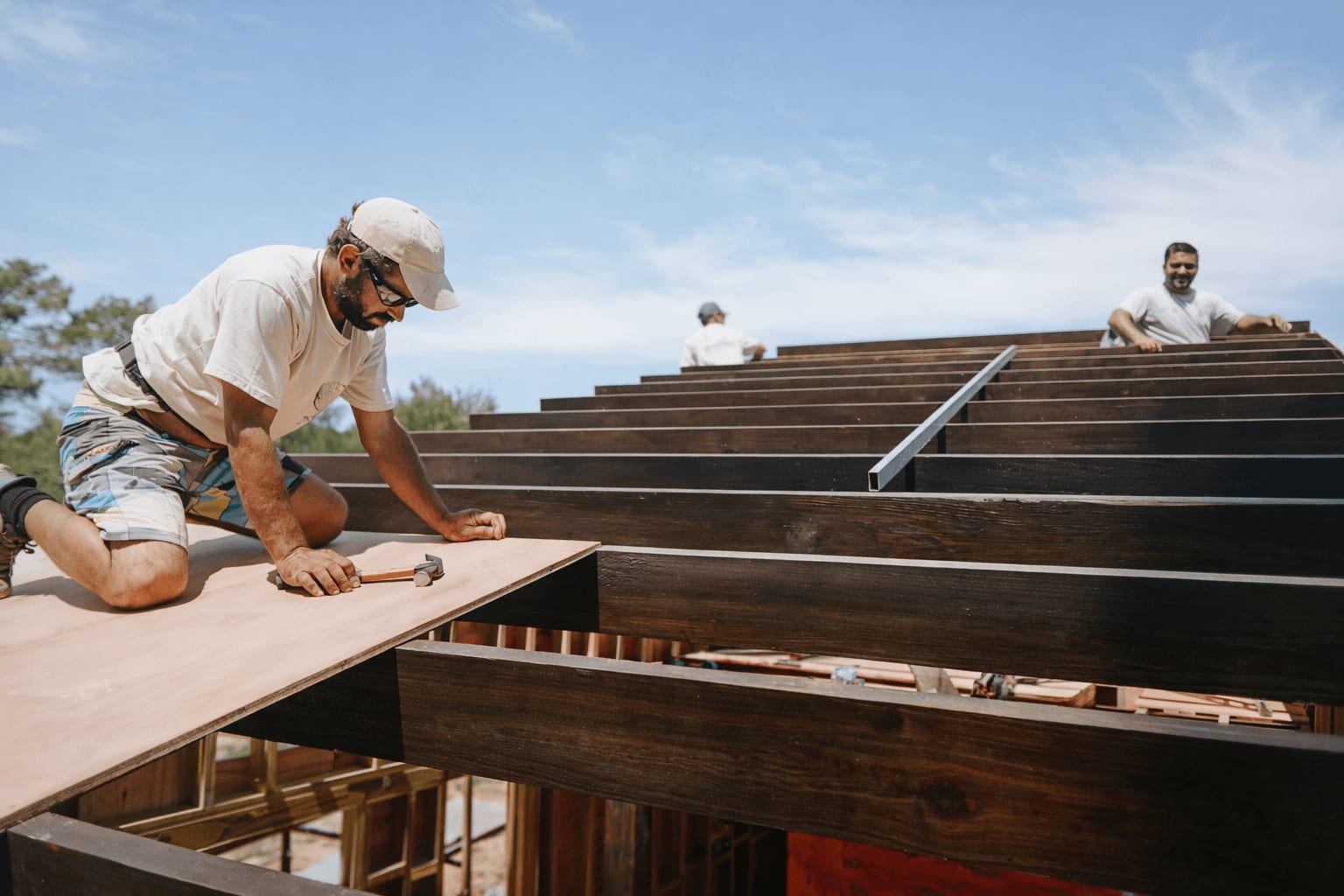 A carpenter nails a roof panel onto dark timber beams, two colleagues in the background.