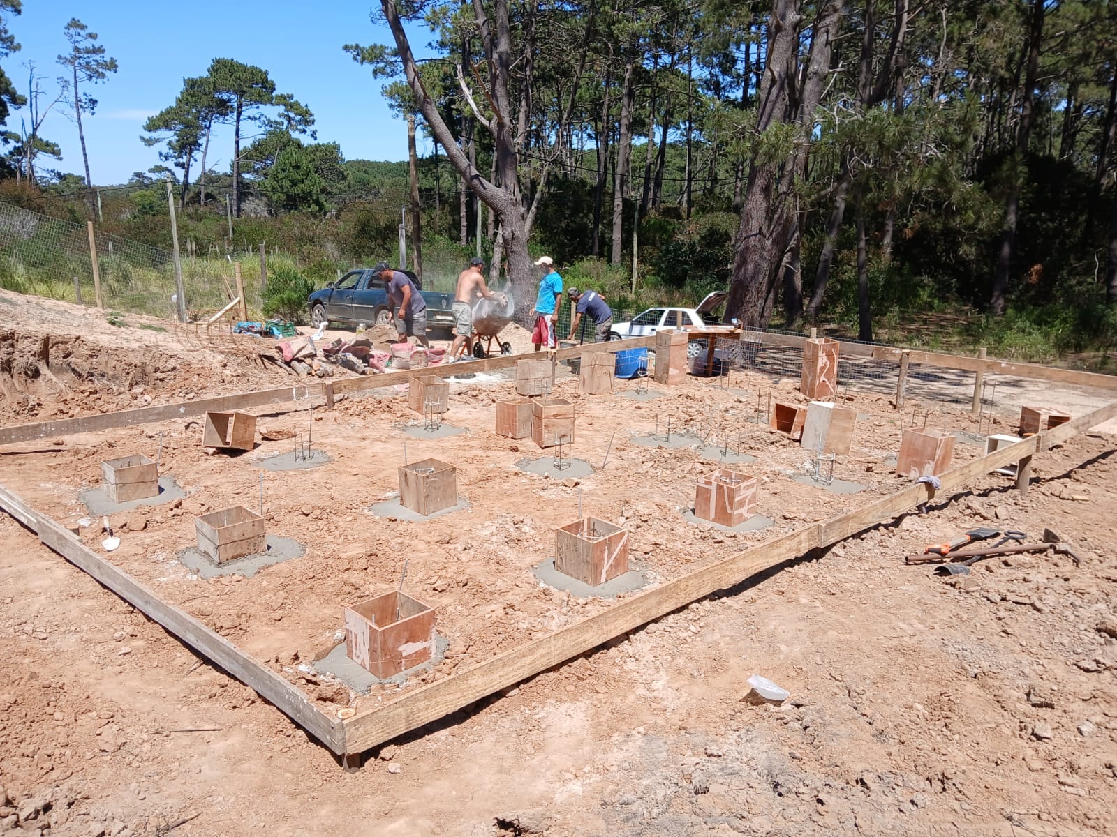 Concrete piers in timber formwork on the staked-out plot, the team at work under the pines in the background.
