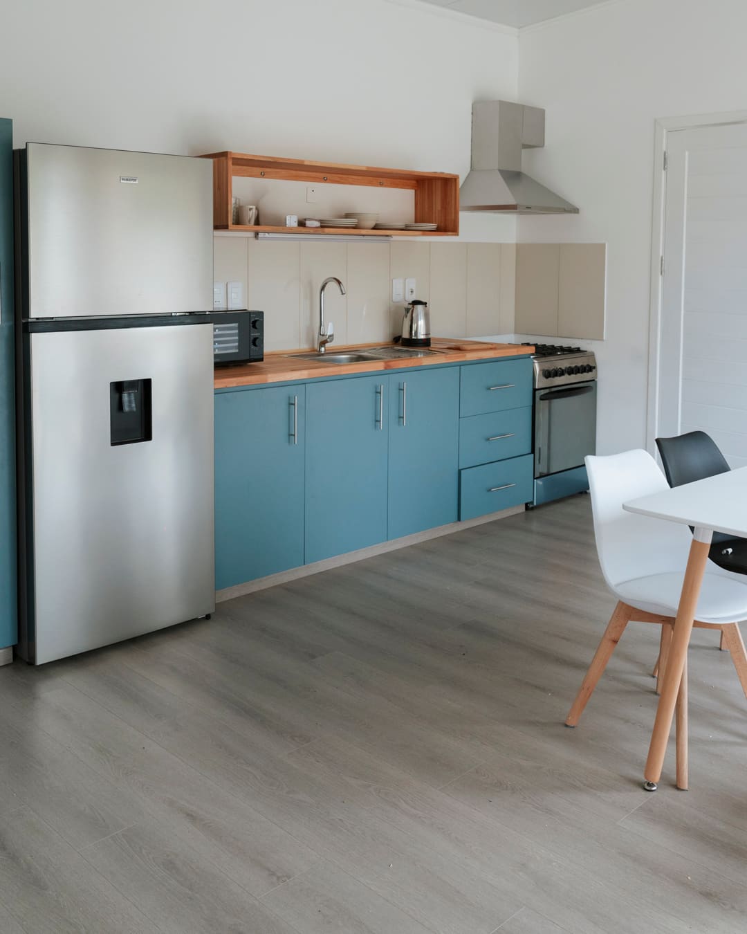 Equipped kitchen with a fridge, microwave and kettle, and a dining table with a white chair at right.