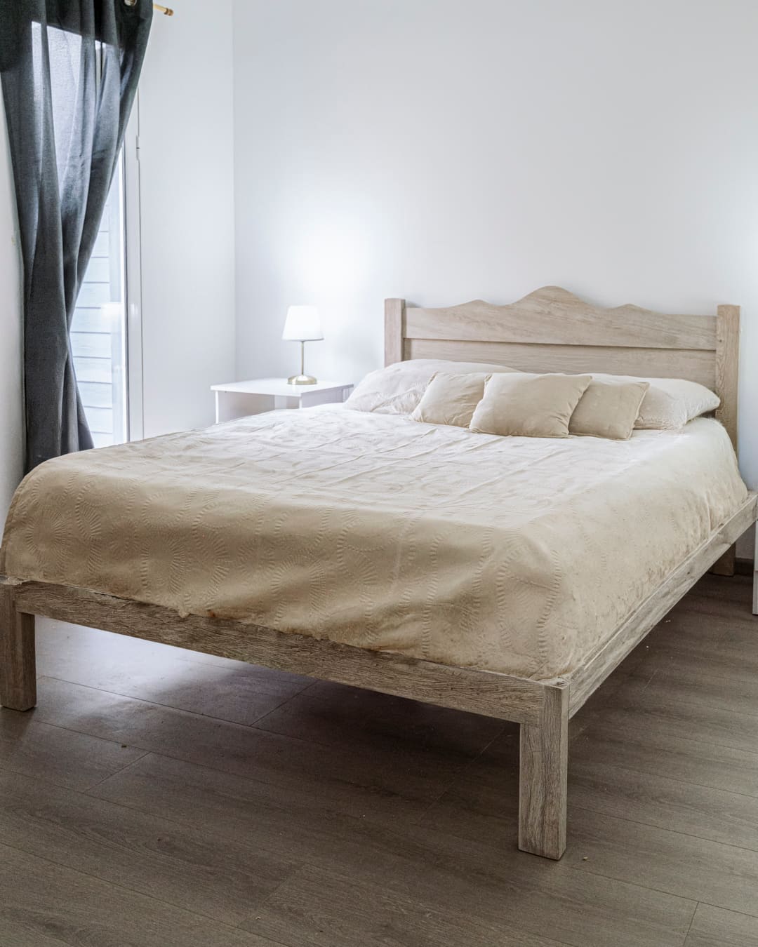 Light wooden double bed with a beige cover and cushions, and a white bedside table with a lamp.