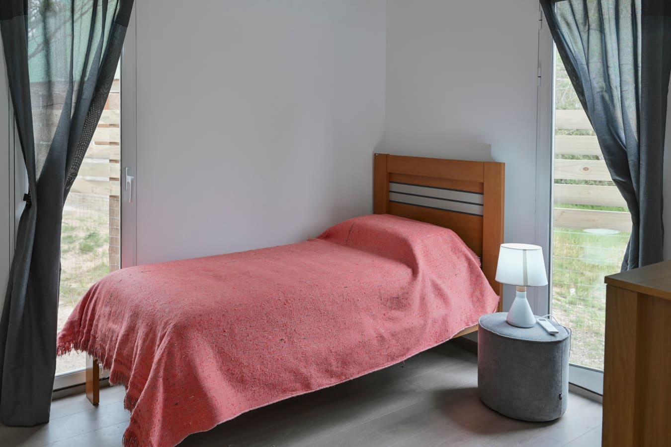Single bed with a red blanket in a corner between two floor-to-ceiling windows with grey curtains.