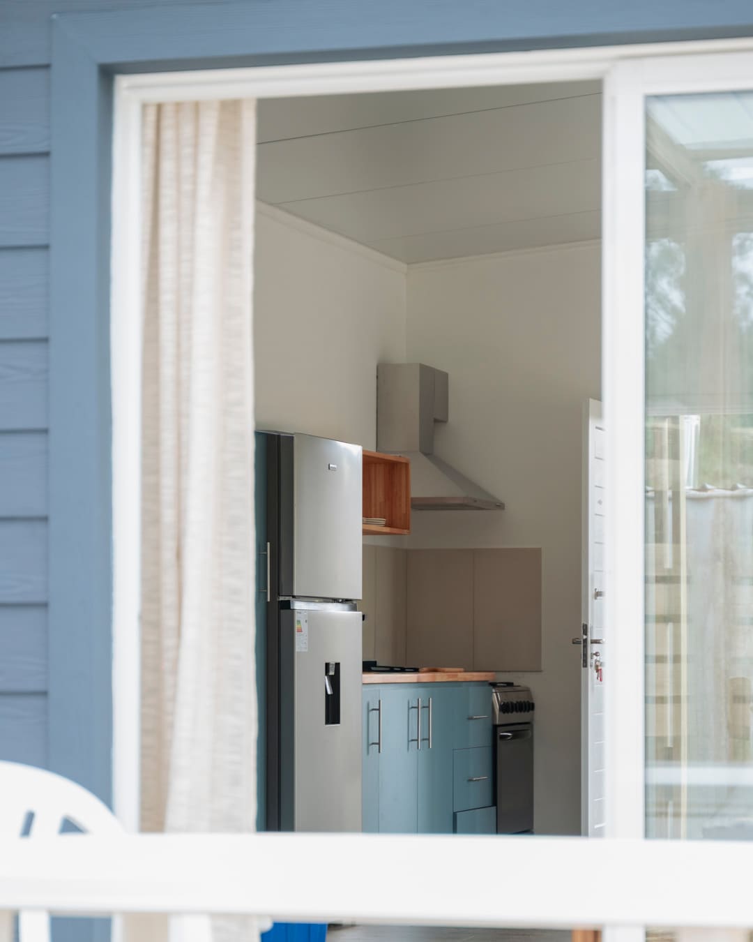 View through the open sliding door and a linen curtain into the kitchen with teal cabinets and a fridge.