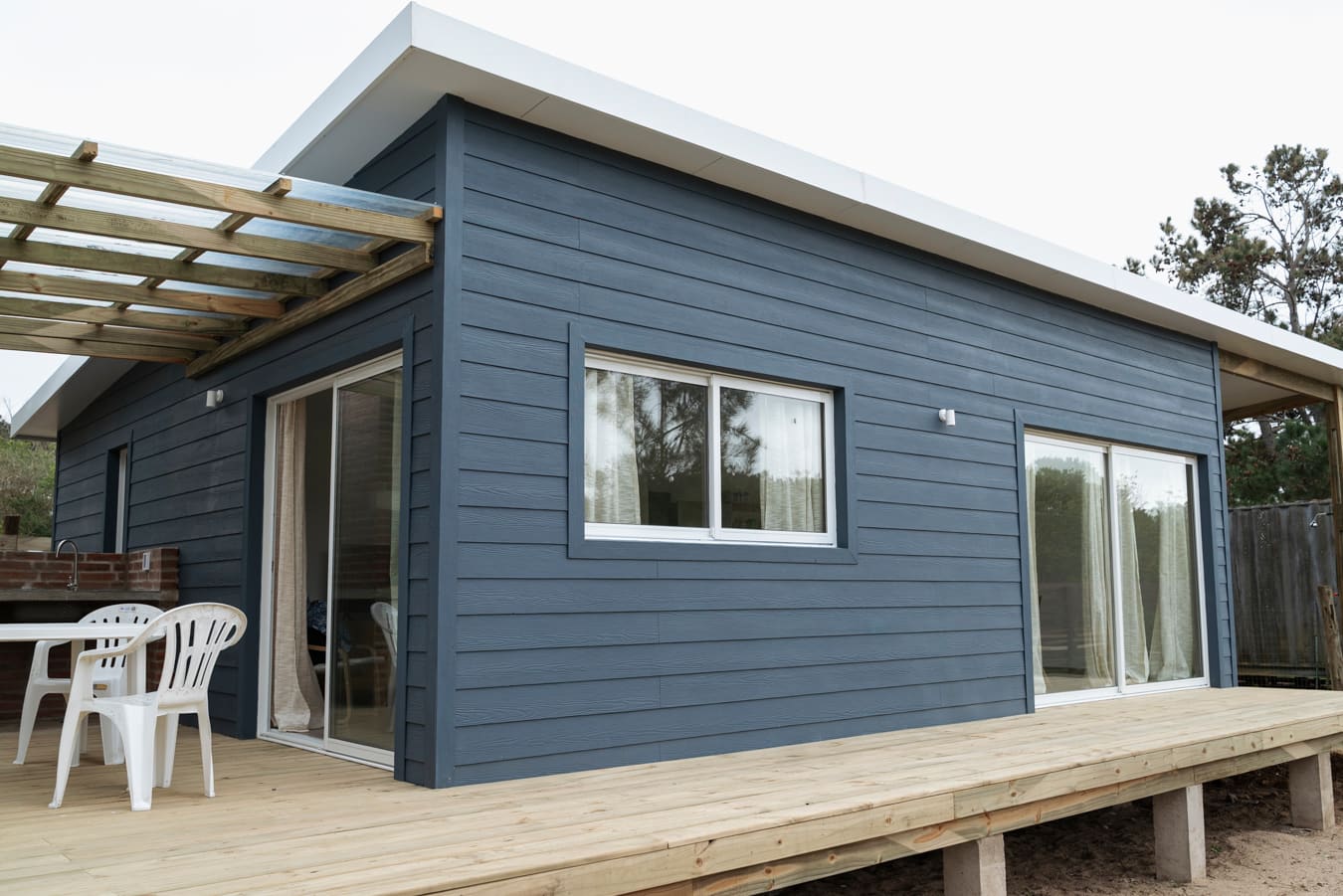 Blue-grey wooden facade with windows and sliding glass doors, a pergola with white plastic chairs at left.