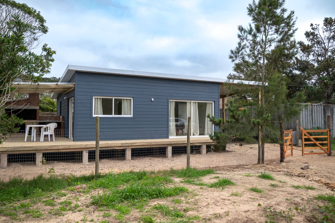 Blue wooden house with a deck and pergola, a wire fence, pine trees and an orange wooden gate in front.