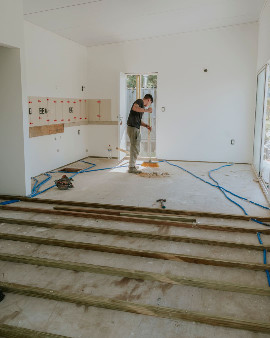 A worker sweeps the bare floor of a bright room, with wooden battens in front and kitchen wall panels behind.