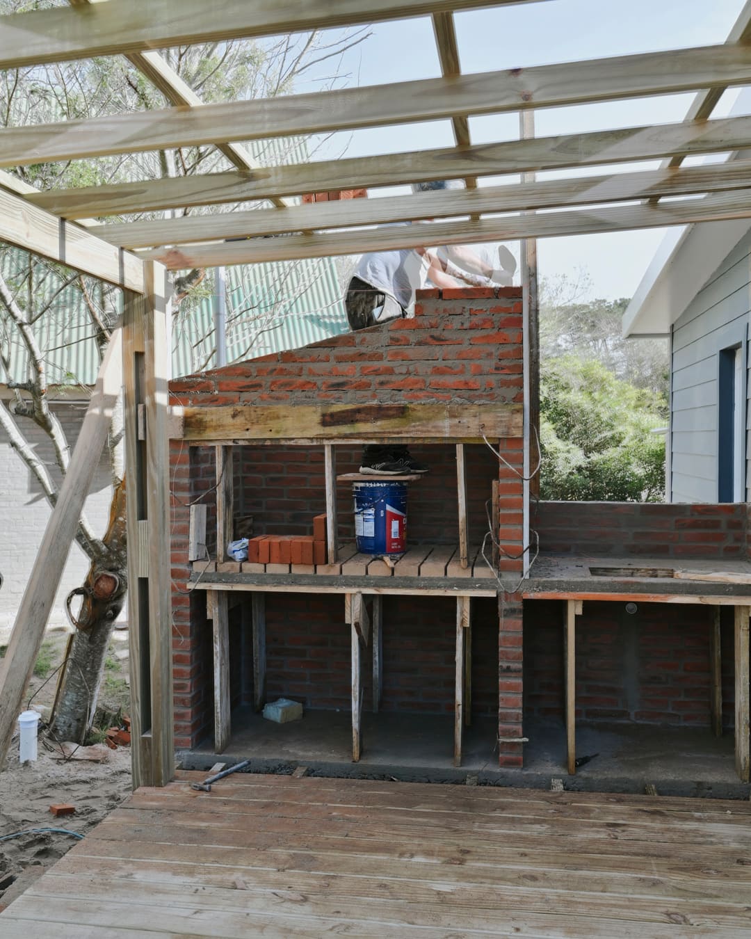Brick barbecue under construction beneath the wooden beams of a pergola, with a timber deck in front.