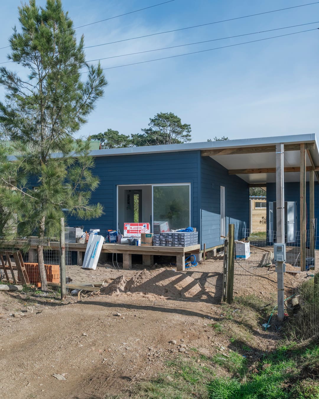 Blue-clad wooden house with a raised deck holding building materials, a pine tree and sandy ground in front.