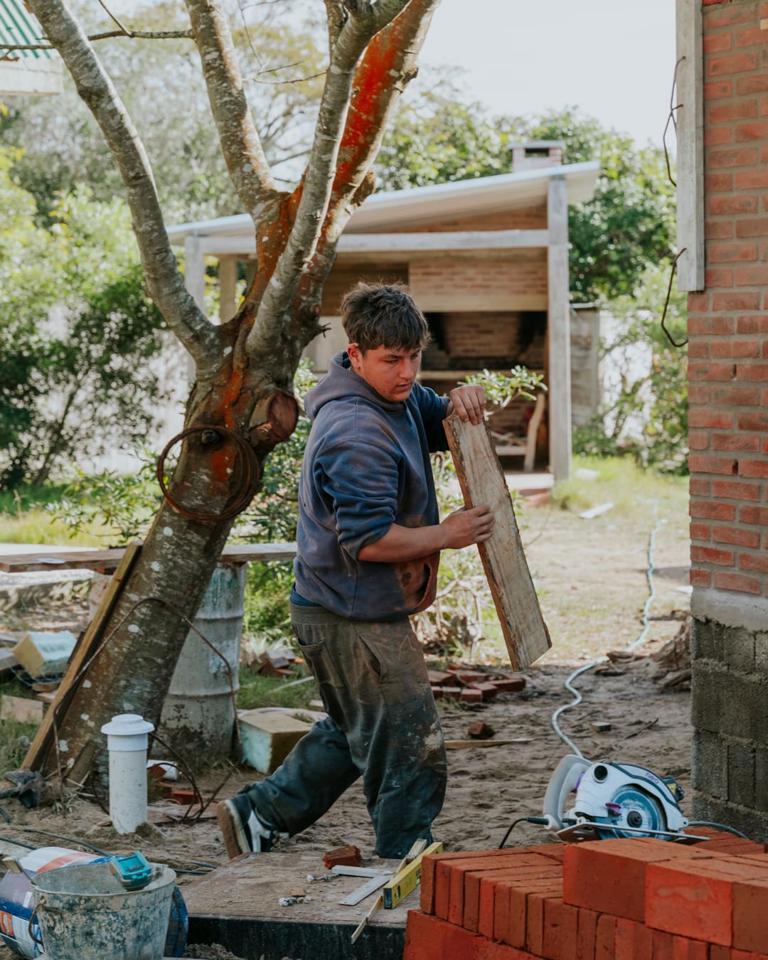 A worker carries a board past a tree, with a stack of bricks and a barbecue under construction behind.
