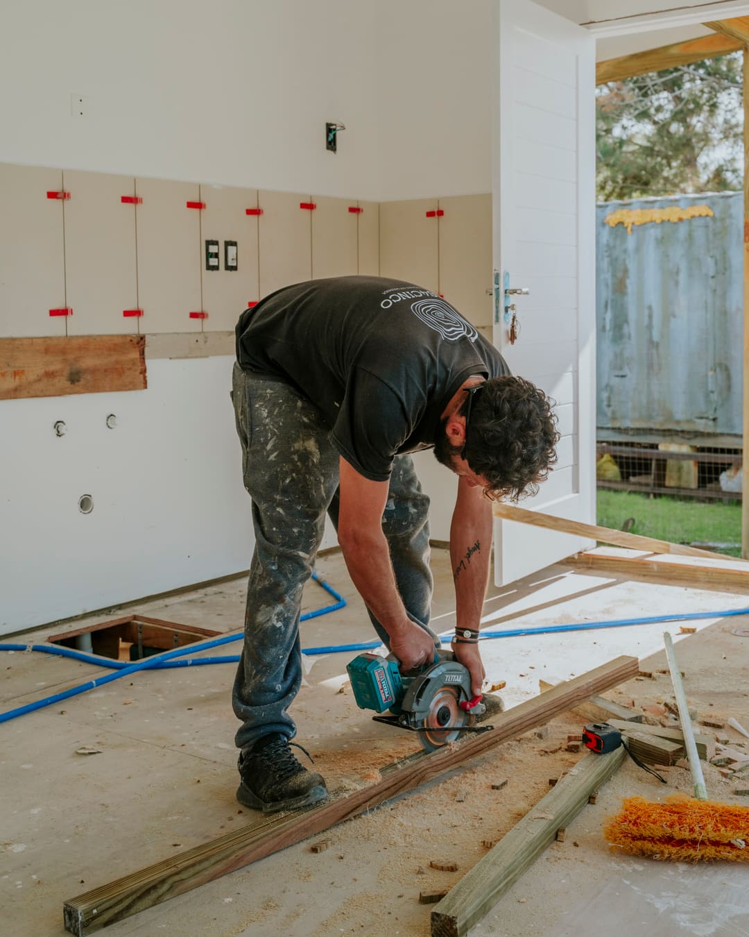 A worker cuts a wooden batten with a circular saw, a wall of backing panels behind him.