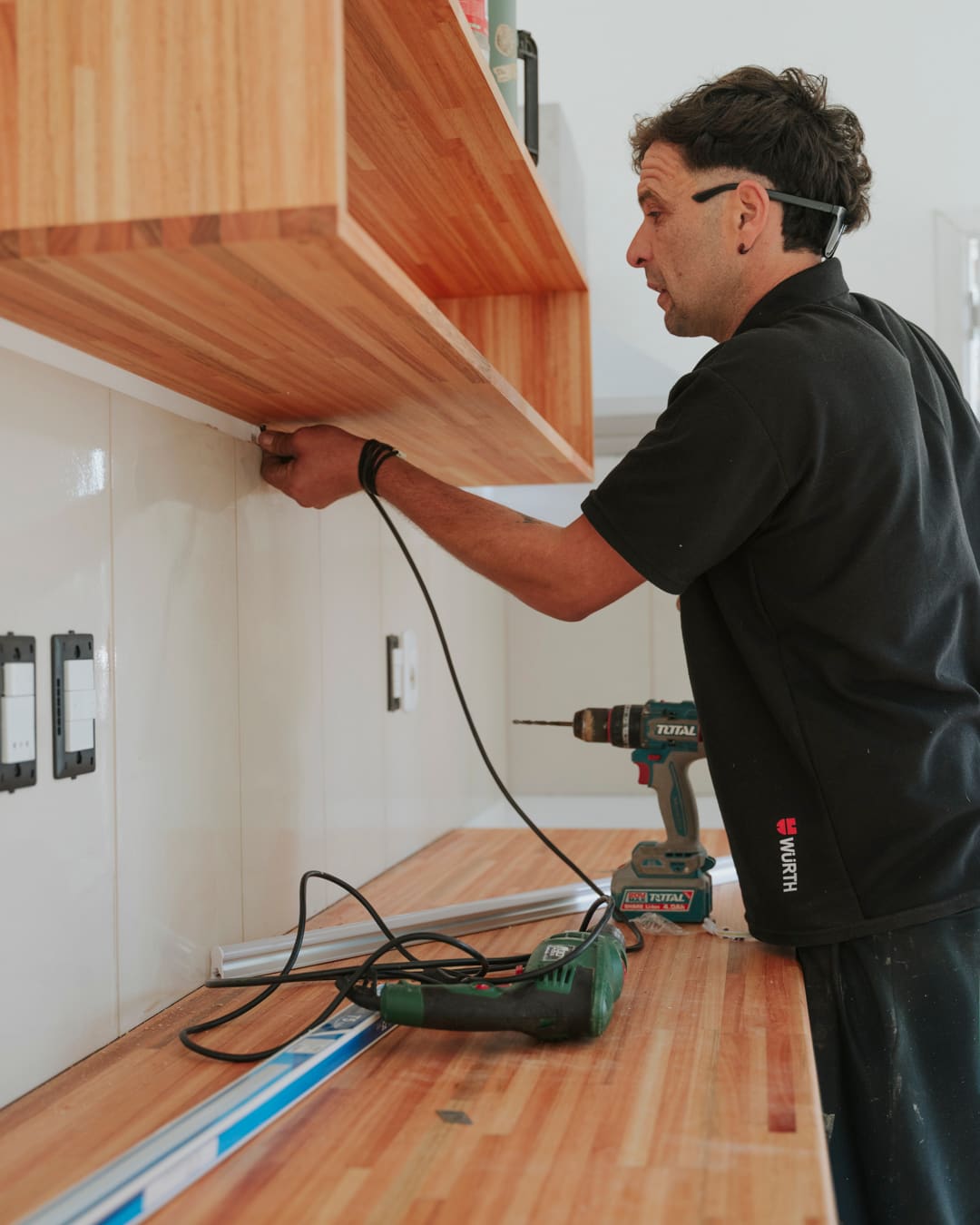 A worker mounts an open wooden shelf above a wooden countertop using a cordless drill.