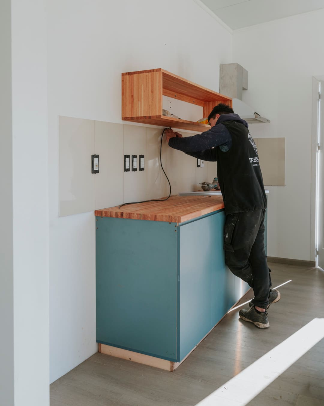 A worker fits the kitchen with teal base cabinets, a wooden countertop and a wooden shelf.