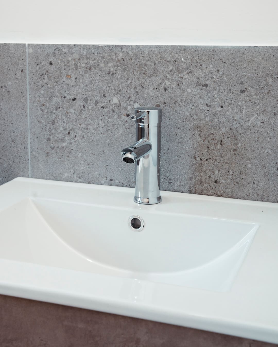 White washbasin with a chrome tap in front of grey stone-look tiles.
