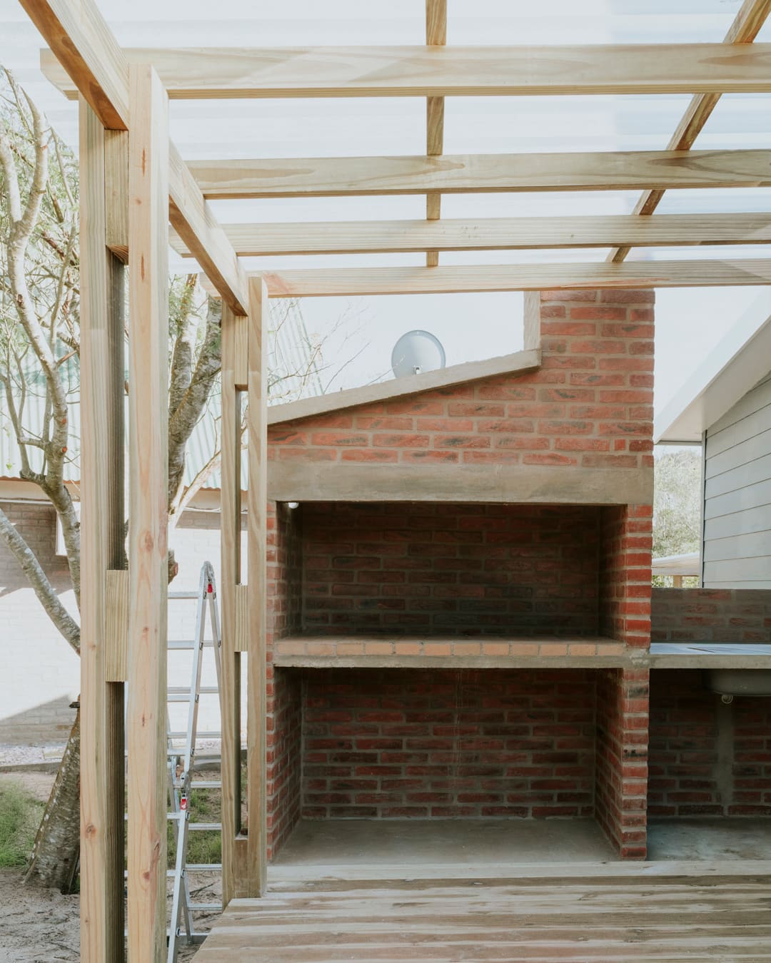 Brick barbecue with a worktop under a wooden pergola, a timber deck in front and a ladder beside it.