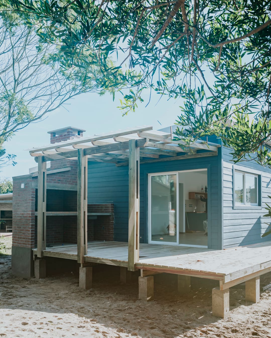 Blue-grey wooden house with a deck, a pergola and a brick barbecue on the left, over pale sand.