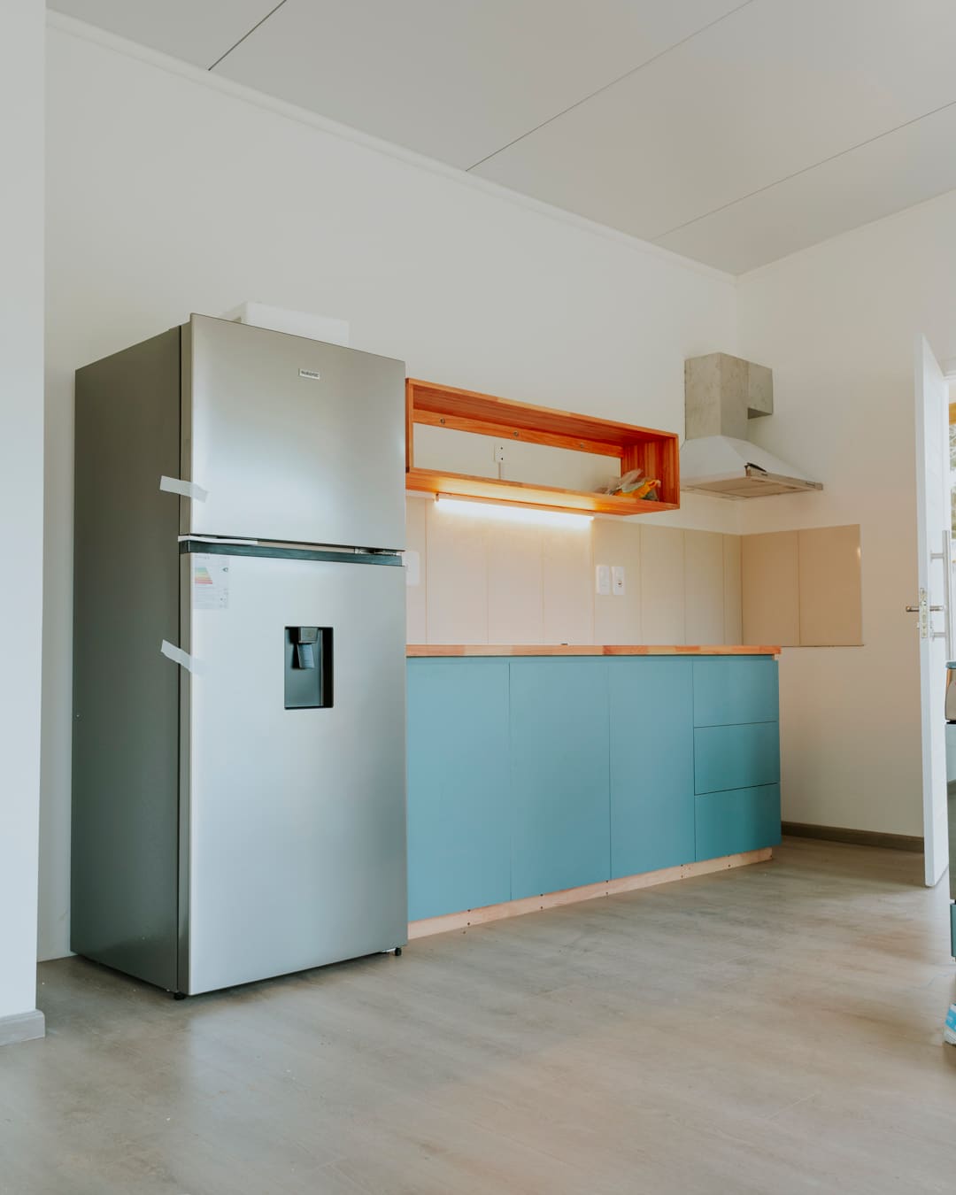 Kitchen run with teal fronts, a wooden shelf, a cooker hood and a stainless steel fridge.