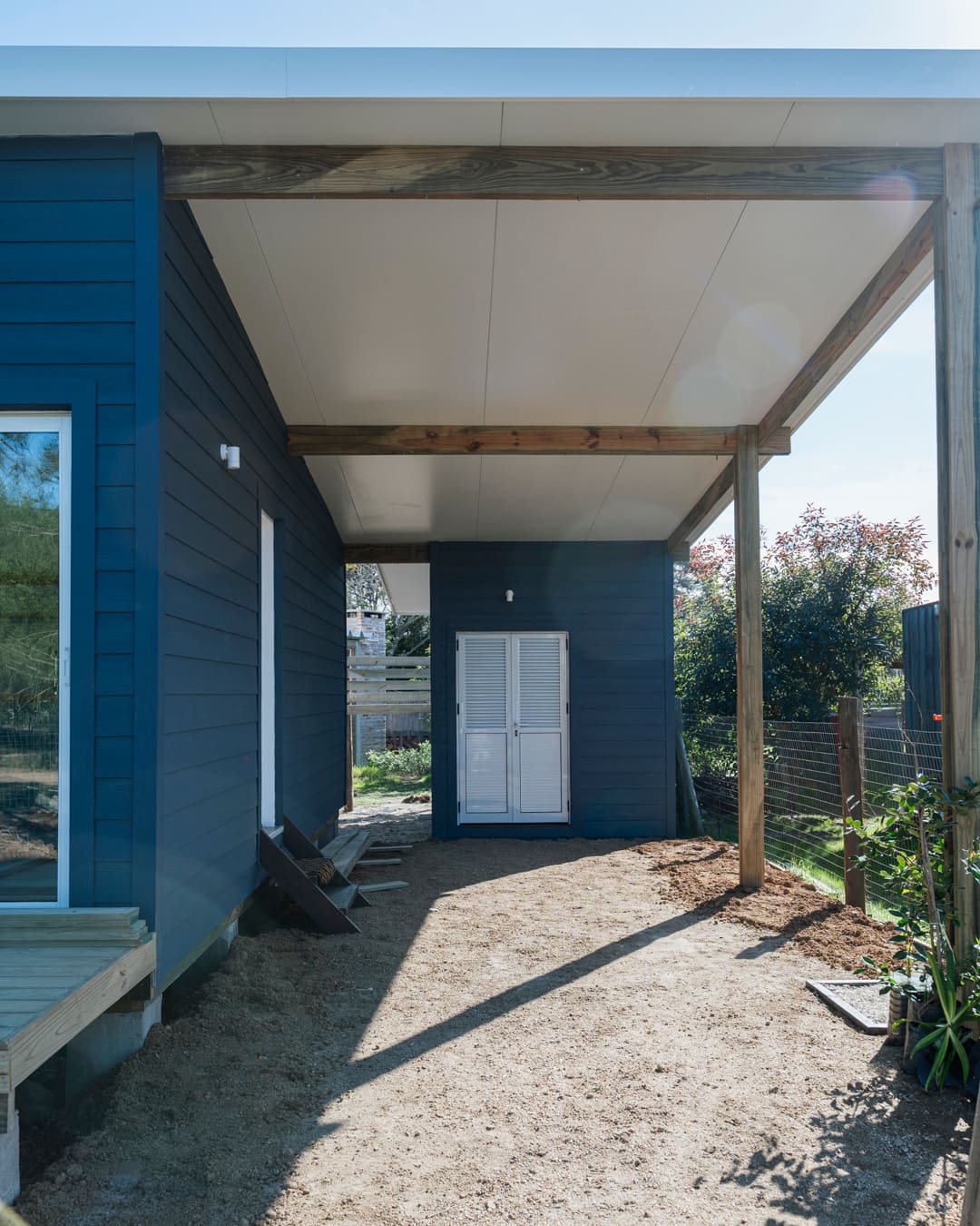Covered passage with timber beams and posts, a white louvered door at the end and the blue facade at left.