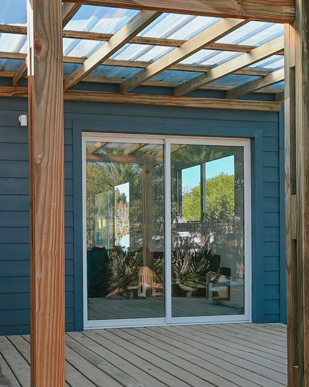 Wooden pergola with a translucent roof over the deck, a sliding glass door in the blue facade behind.